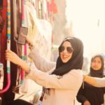 Woman in hijab and sunglasses climbing a decorated bus on a sunny day.